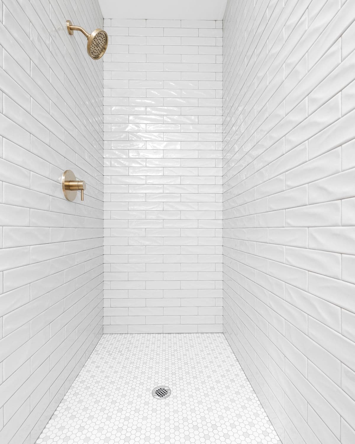 A finished walk-in shower tiled in white subway tile with warm brass fixtures and a penny-tile floor.